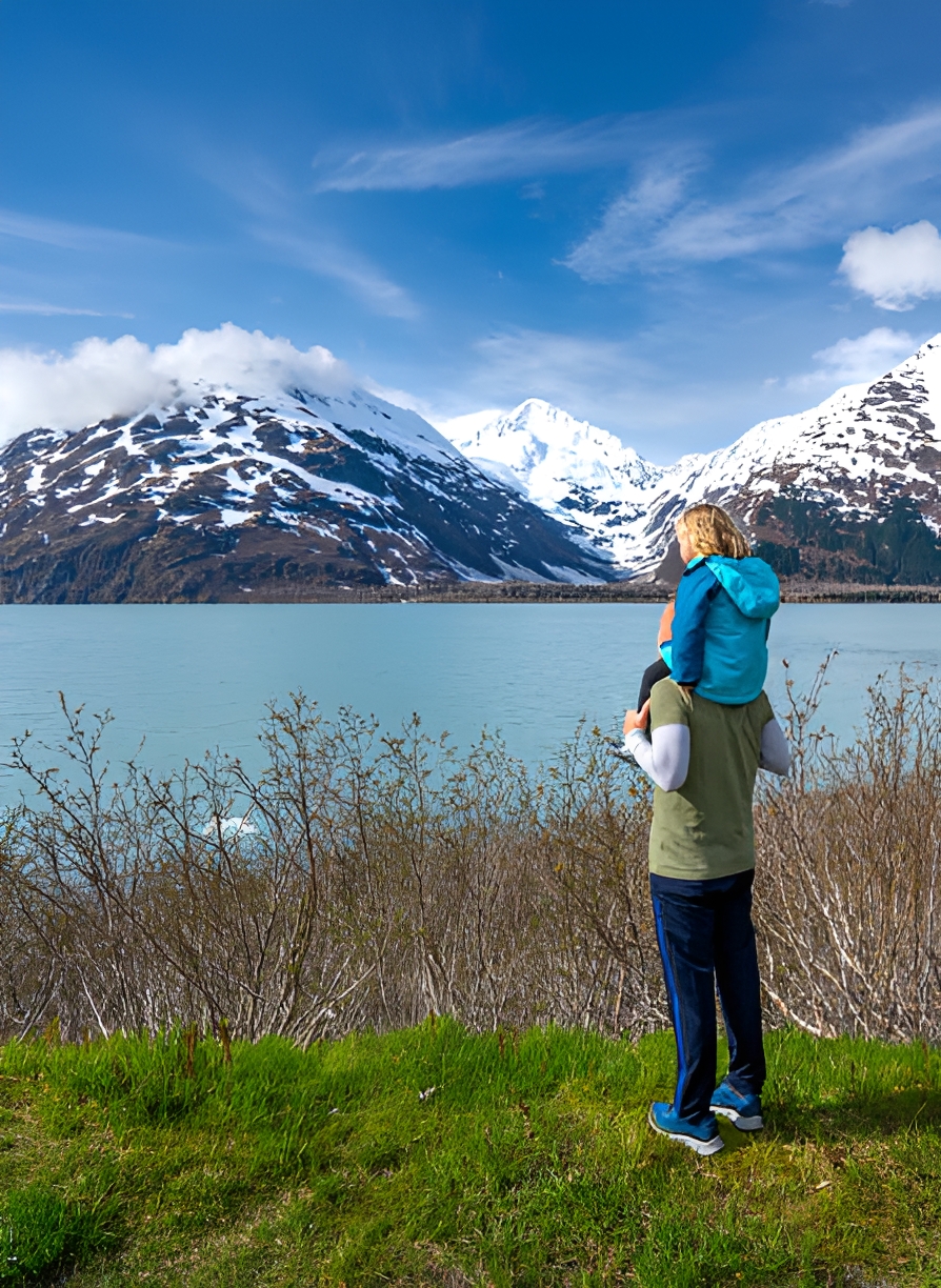 Child on shoulders near snowy mountains and lake.